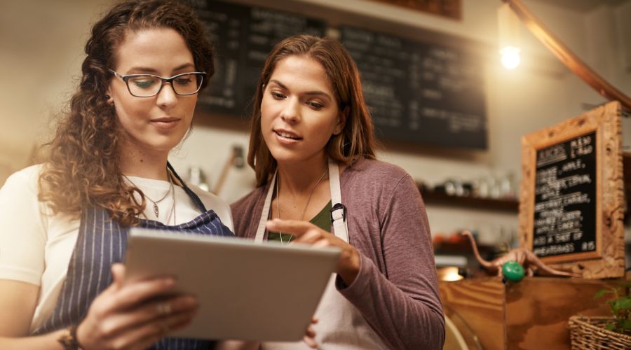 two women wearing aprons looking at a tablet while standing in a bakery